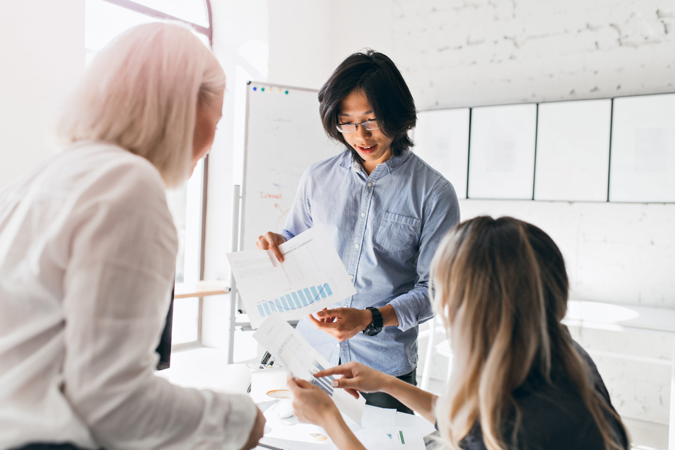 Asian male office worker with wristwatch holding documents with diagrams while talking with female colleagues. Indoor portrait of young executive in glasses discussing new goals.. Asian male office worker with wristwatch holding documents with diagrams while talking with female colleagues. Indoor portrait of young executive in glasses discussing new goals.